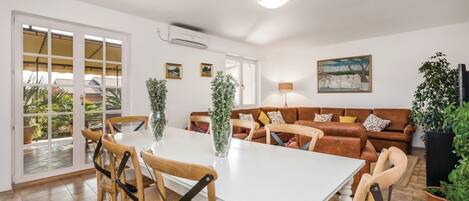 A spacious living area with a white dining table, wooden chairs, a brown sofa, and a large window overlooking a garden.