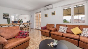 Sala de estar con sofás de cuero marrón, una mesa de comedor y sillas. Hay una mesa de centro negra con objetos decorativos. La habitación tiene paredes blancas, piso de baldosas y ventanas grandes con persianas.