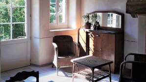 Una habitación con un ventanal grande, una silla de mimbre, una mesita y un aparador con adornos.