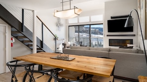 Un salon moderne avec une table à manger en bois, des chaises en métal noir, un canapé gris et une grande fenêtre donnant sur la ville.