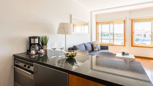 Eine moderne Küche mit Einbauofen, Kaffeemaschine und Weingläsern auf der Arbeitsfläche. Ein Essbereich mit Sofa und Lampe ist ebenfalls vorhanden.
