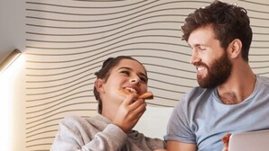 Una pareja en un dormitorio con una computadora portátil, una caja de pizza y un control remoto sobre la mesita de noche.