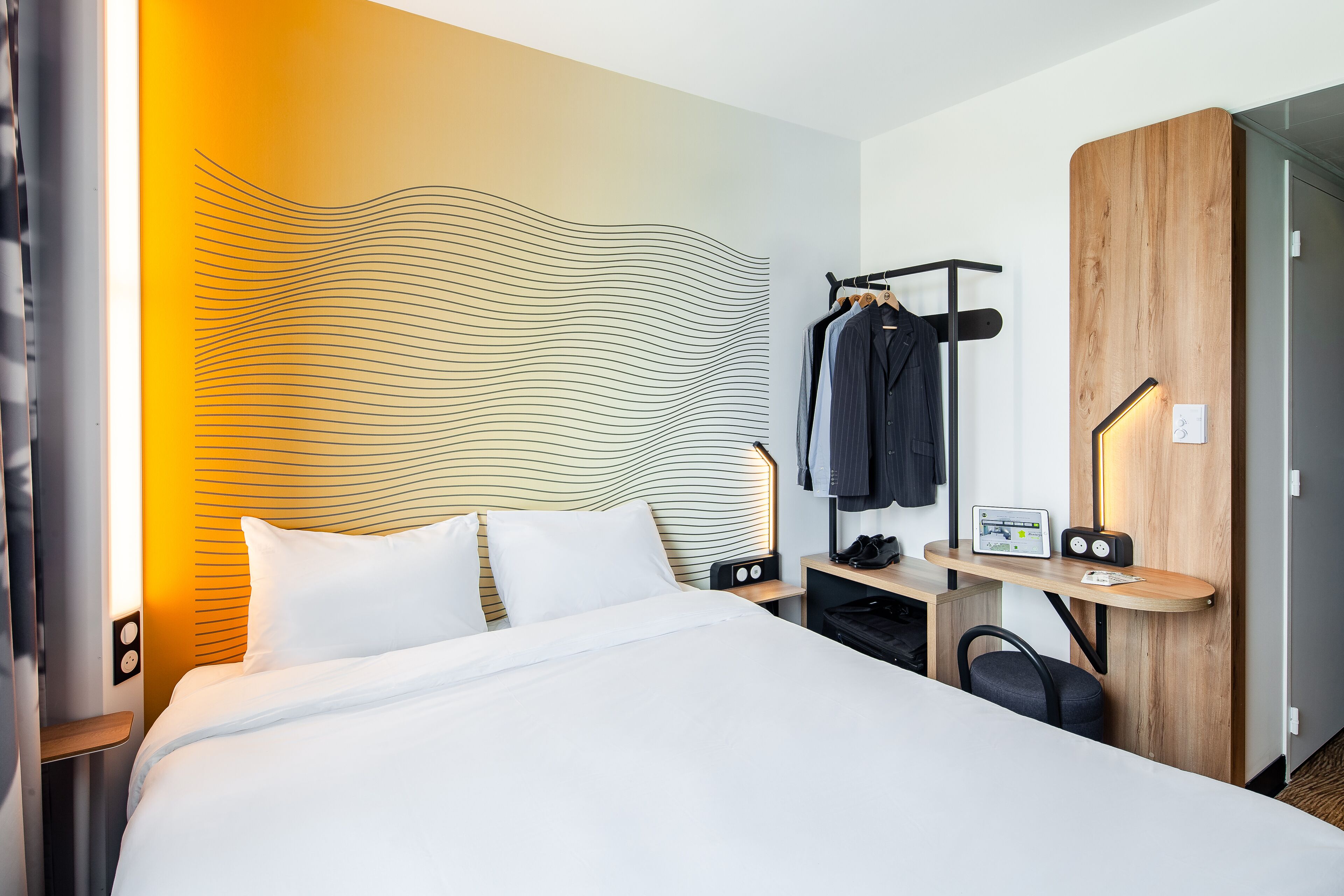 A modern hotel room with a large bed, a wooden headboard, a desk with a chair, and a coat rack.
