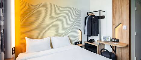 A modern hotel room with a large bed, a wooden headboard, a desk with a chair, and a coat rack.
