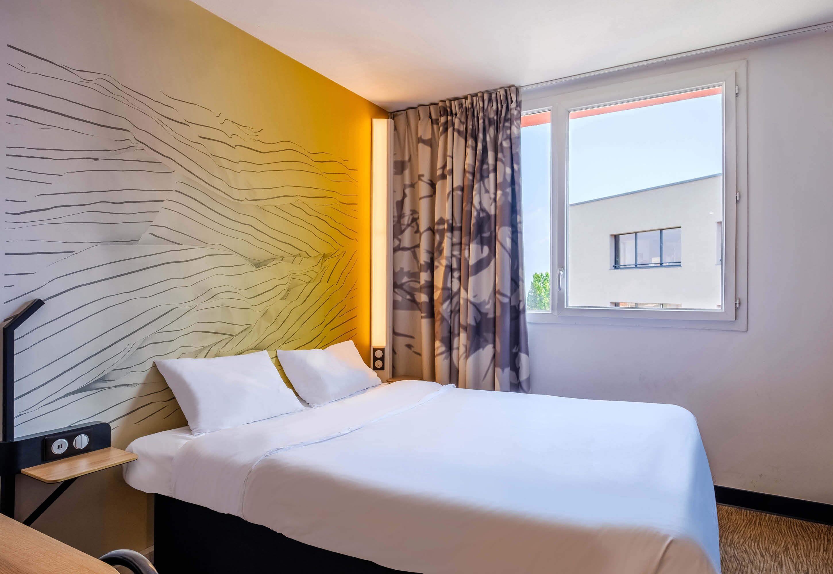 Een hotelkamer met een bed, nachtkastje met lamp, wand met bladmotief en raam met gordijnen.
