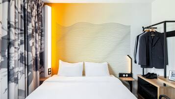 Ein Hotelzimmer mit einem ordentlich bezogenem Bett, einem Nachttisch mit einer Lampe, einem Kleiderhaken mit einem Anzug und einer Wand mit einem gewellten Muster.