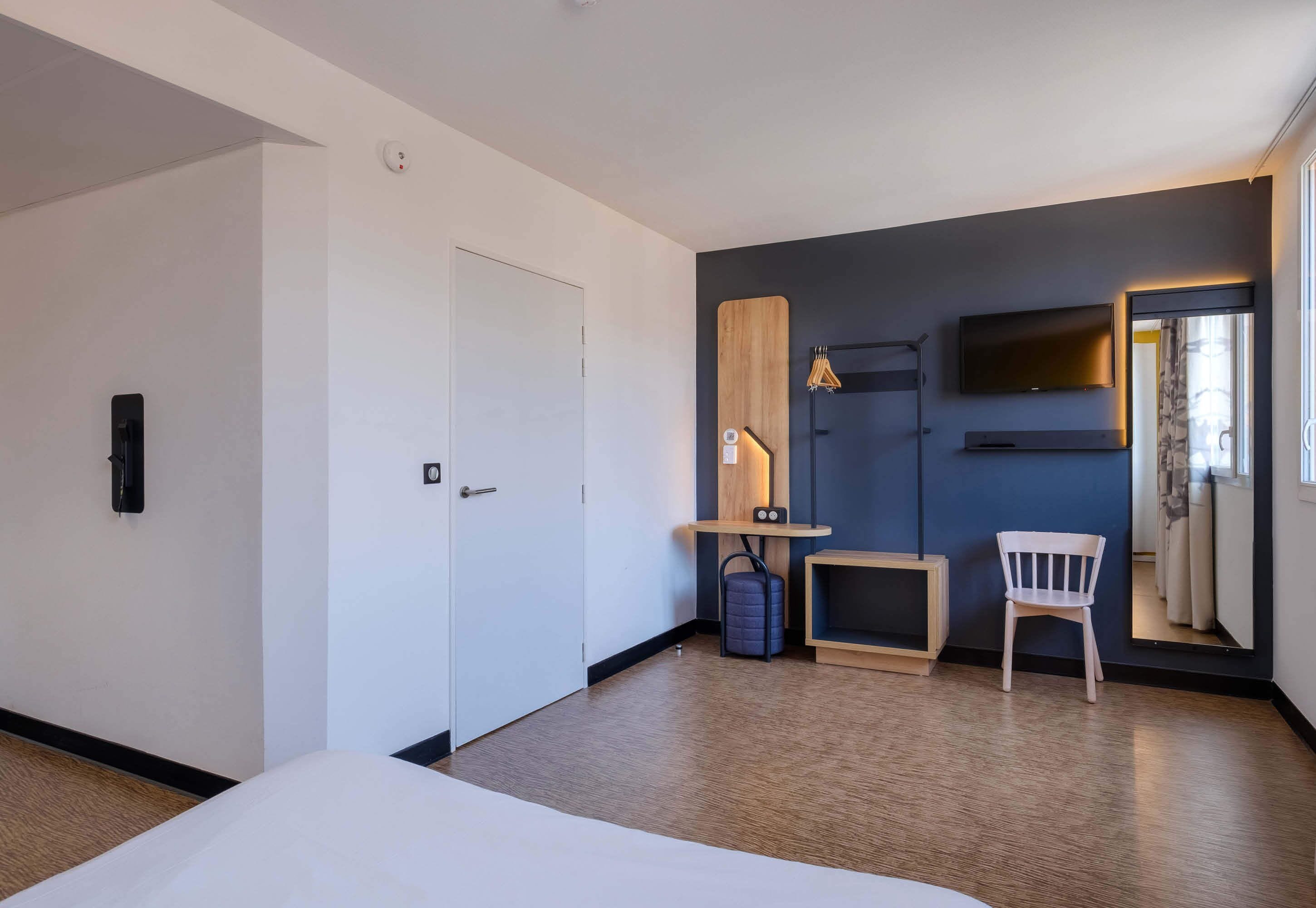 A modern hotel room with a bed, a desk, a chair, a TV, and a window with curtains.