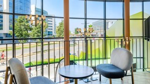 Un balcón con dos sillas y una mesita, ventanas amplias con vista a la ciudad y un edificio moderno al fondo.
