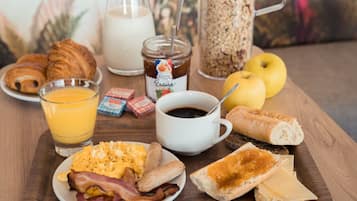 A breakfast table with croissants, scrambled eggs, bacon, cheese, toast, a glass of orange juice, a cup of coffee, a jar of jam, and a whole apple.