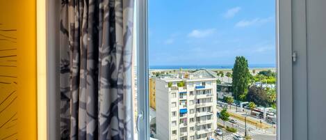 A room with a large window overlooking a cityscape, including a building with balconies and a view of the sea.