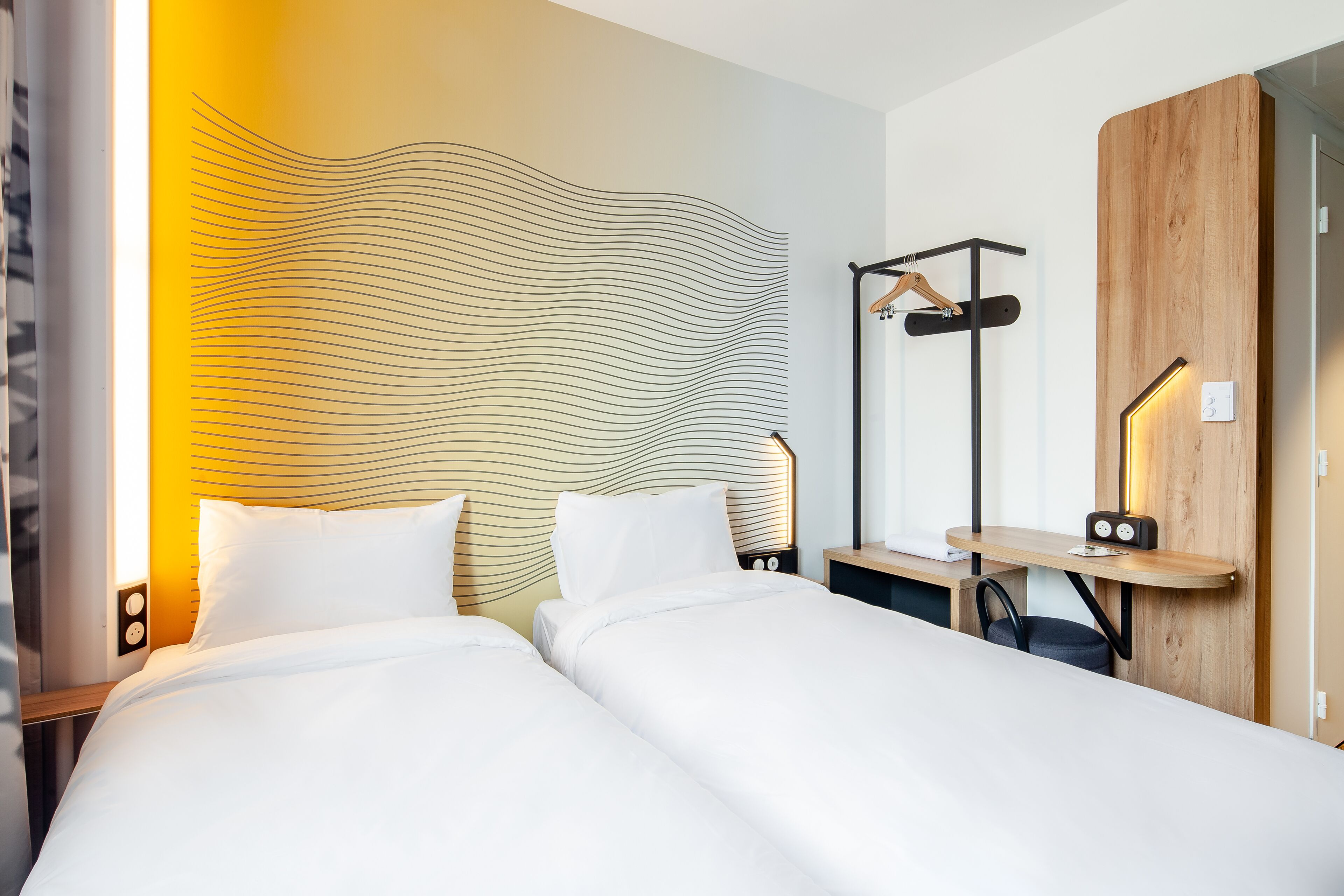 A hotel room with two beds, a wooden headboard, a bedside table with a lamp, and a wooden wardrobe.