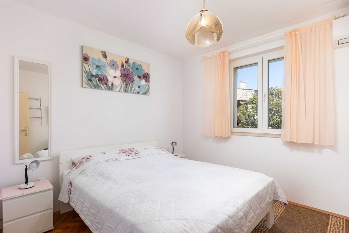 Un dormitorio con cama, mesita de noche con lámpara, espejo, ventana con cortinas y un cuadro con motivos florales en la pared.
