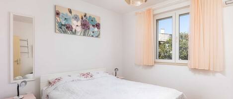 Quarto com cama, mesa de cabeceira com abajur, espelho, janela com cortinas e um quadro floral na parede.