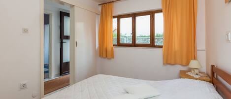 Un dormitorio con cama, dos almohadas, una mesita de noche con lámpara, una ventana con cortinas y una puerta que da a otra habitación.