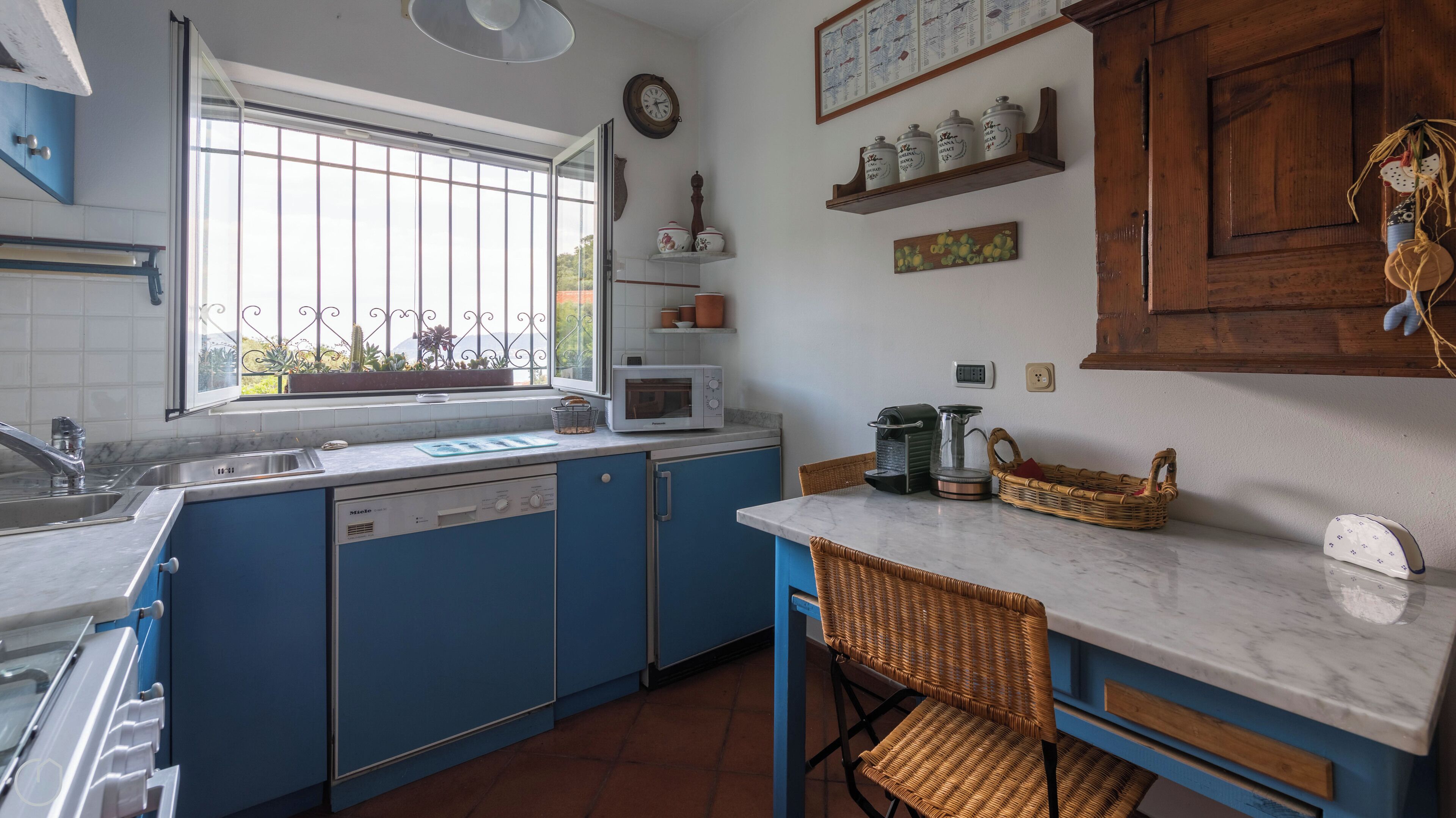A kitchen with blue cabinets, a white countertop, a window with a view, a microwave, a coffee maker, and a wicker chair.