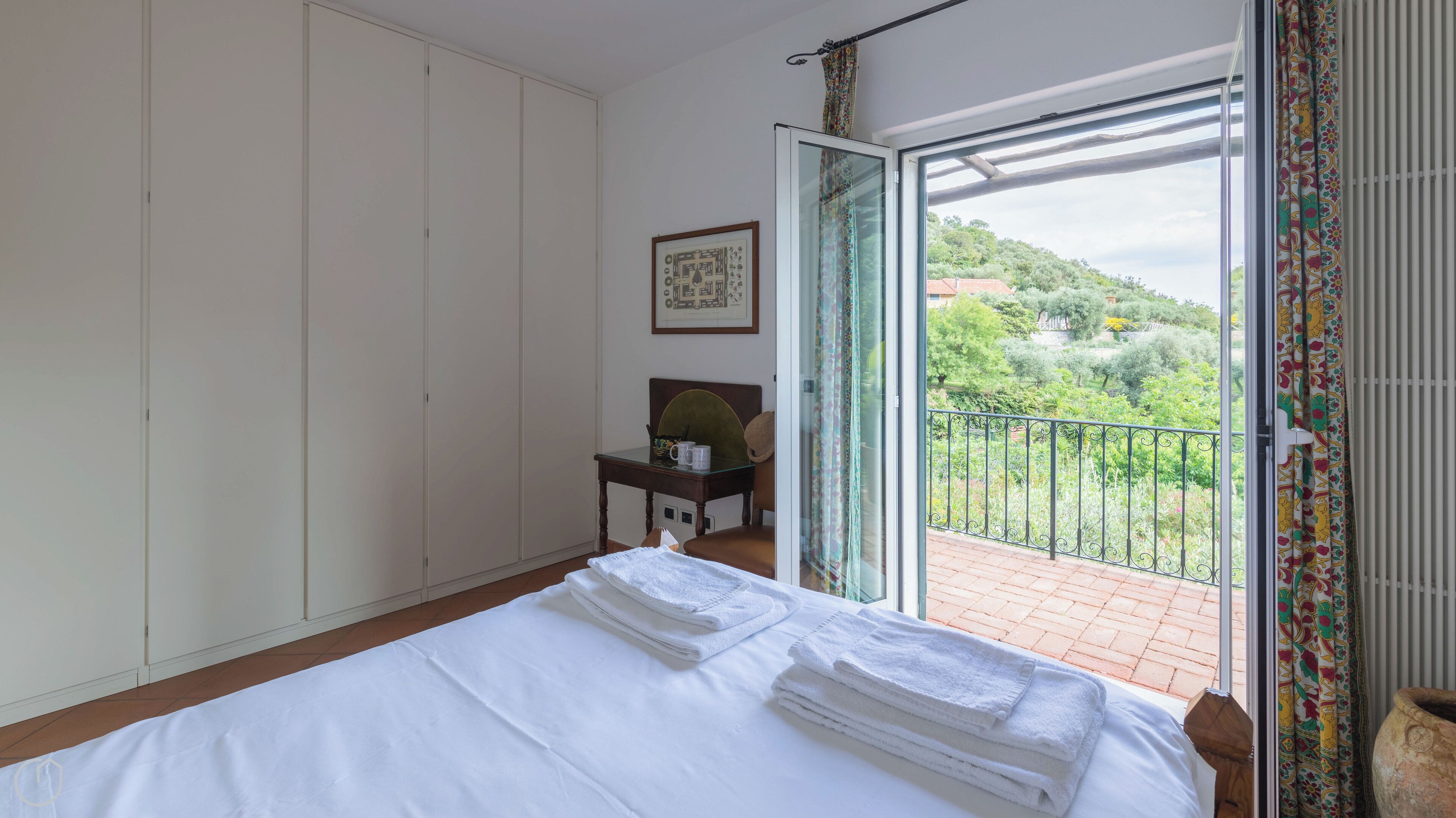 A hotel room with a bed, a small table with a hat and mug, a framed picture on the wall, and a balcony with a view of greenery.