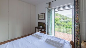 Hotelkamer met een bed, een klein tafeltje met een hoed en mok, een lijst met foto aan de muur en een balkon met uitzicht op groen.