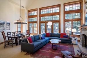 A spacious living room with a large sectional sofa, a dining table with chairs, and large windows offering a view of the outdoors.