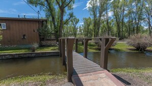 A wooden bridge over a calm river, surrounded by trees and a wooden building with a stone foundation. - River House on the River (Gunnison)