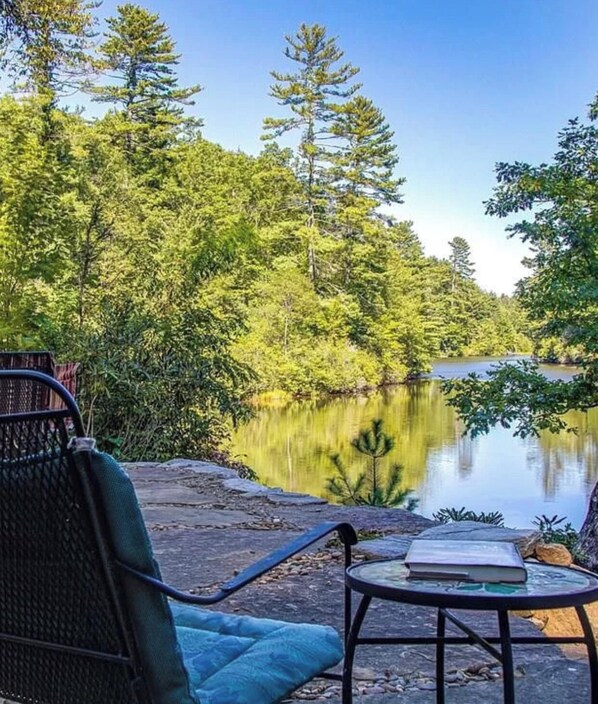 A serene lakeside setting with a chair and a table, surrounded by dense forest and a calm body of water. - Lakefront 3 bedroom cabin sleeps 10, 8 minute drive to Gorges State Park (Lake Toxaway)