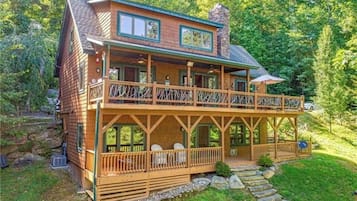 A two-story log cabin with a balcony, stone steps leading to the entrance, and a surrounding wooded area.