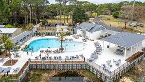 An aerial view of a resort with a large swimming pool surrounded by lounge chairs and umbrellas, a covered patio area, and a dock extending into a body of water.