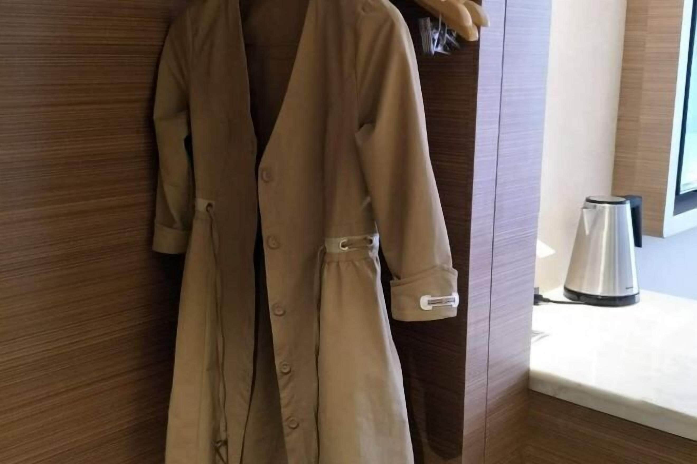 A beige coat hanging on a wooden wall in a room with a window and a kettle on a table.