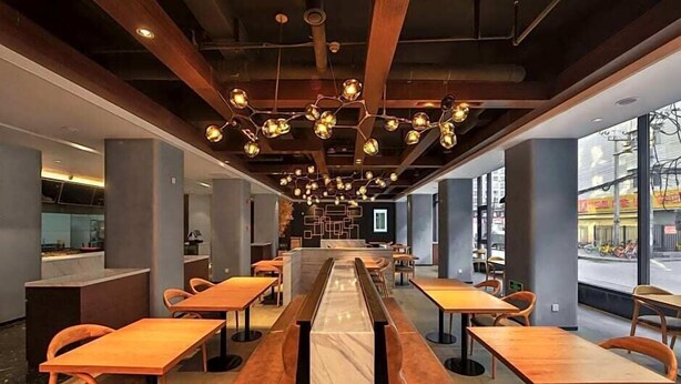 Ein modernes Restaurant mit langen Gemeinschaftstischen und Sitzecken, einer zentralen Bar und großen Fenstern mit Blick auf die Straße.
