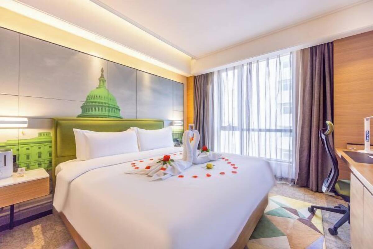 A hotel room with a bed, a desk, a chair, a lamp, a window with curtains, and a wall with a green dome structure.