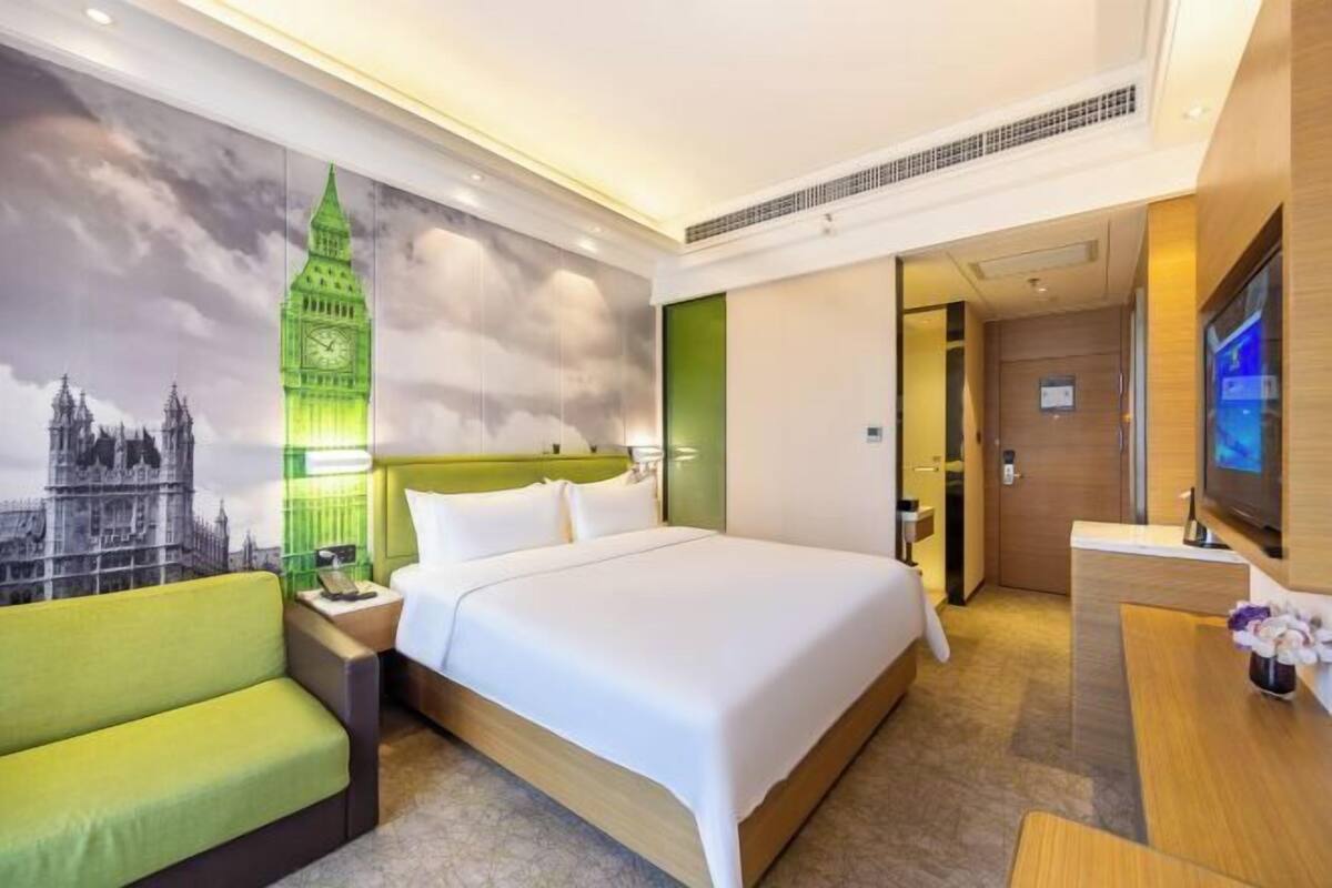 A hotel room with a large bed, a green armchair, a flat-screen TV, and a wall mural of a famous clock tower.