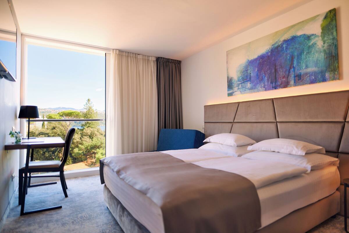A hotel room with a large bed, a desk with a chair, a painting on the wall, and a view of the outdoors.