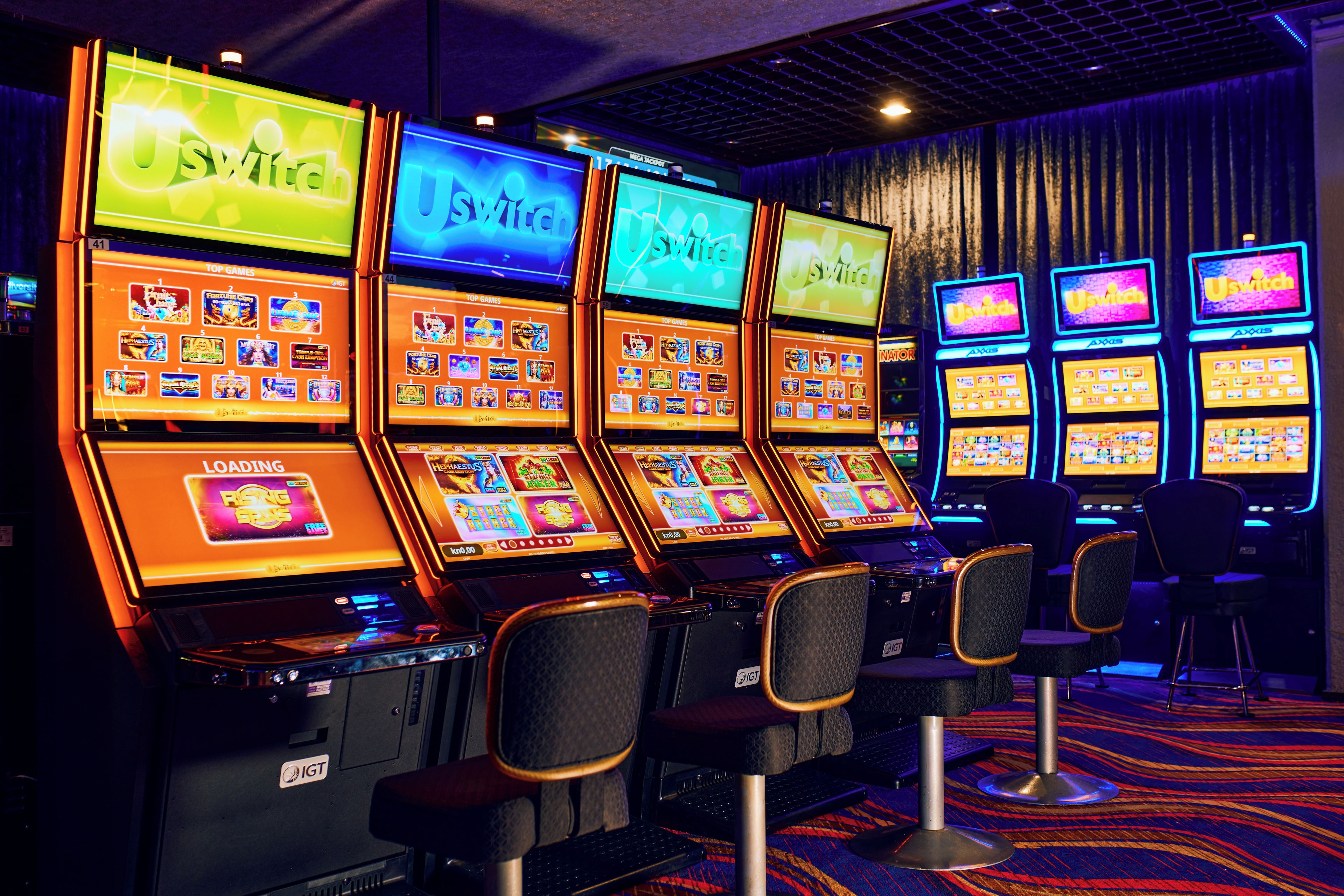 A row of slot machines with illuminated screens and chairs in front.