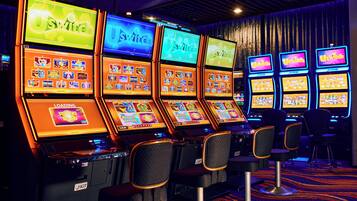A row of slot machines with illuminated screens and chairs in front.