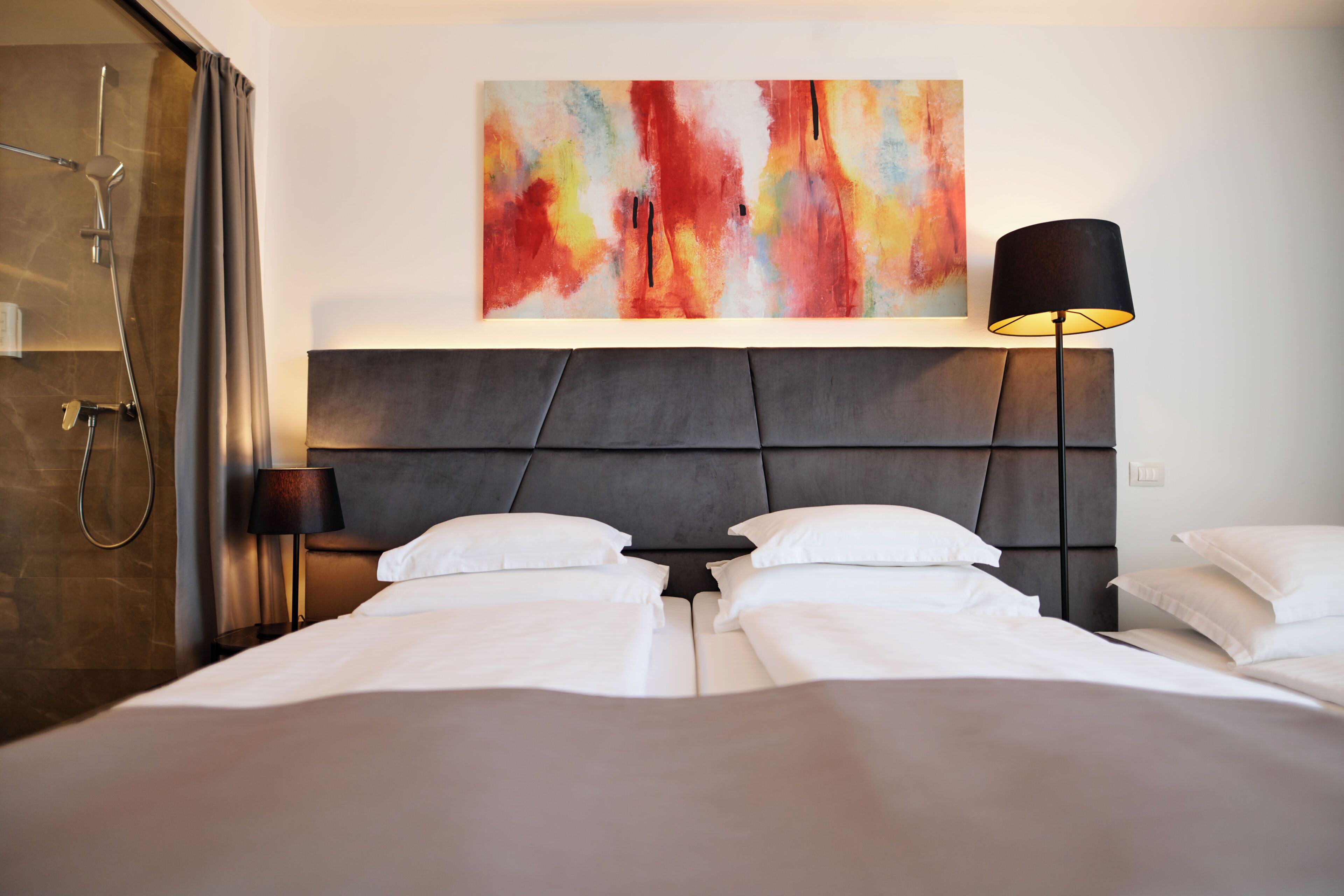 A hotel room with a bed, a shower, a wall-mounted painting, and a floor lamp.