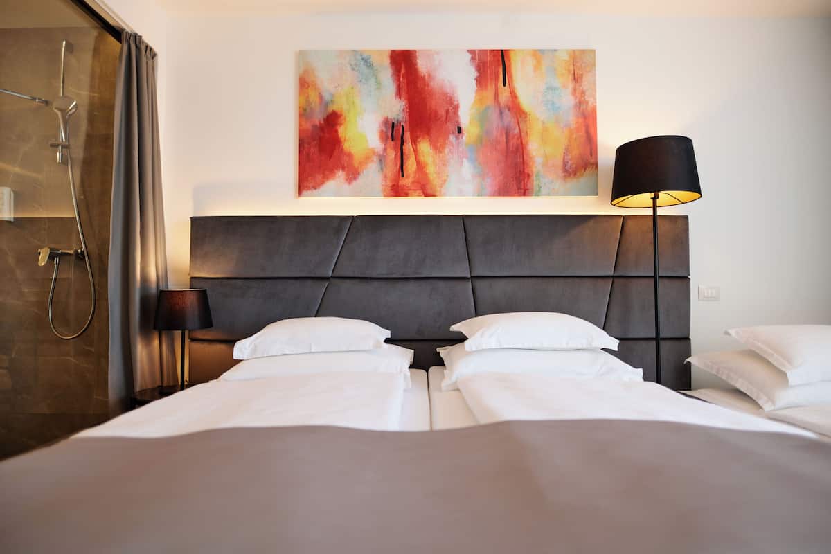 A hotel room with a bed, a shower, a wall-mounted painting, and a floor lamp.