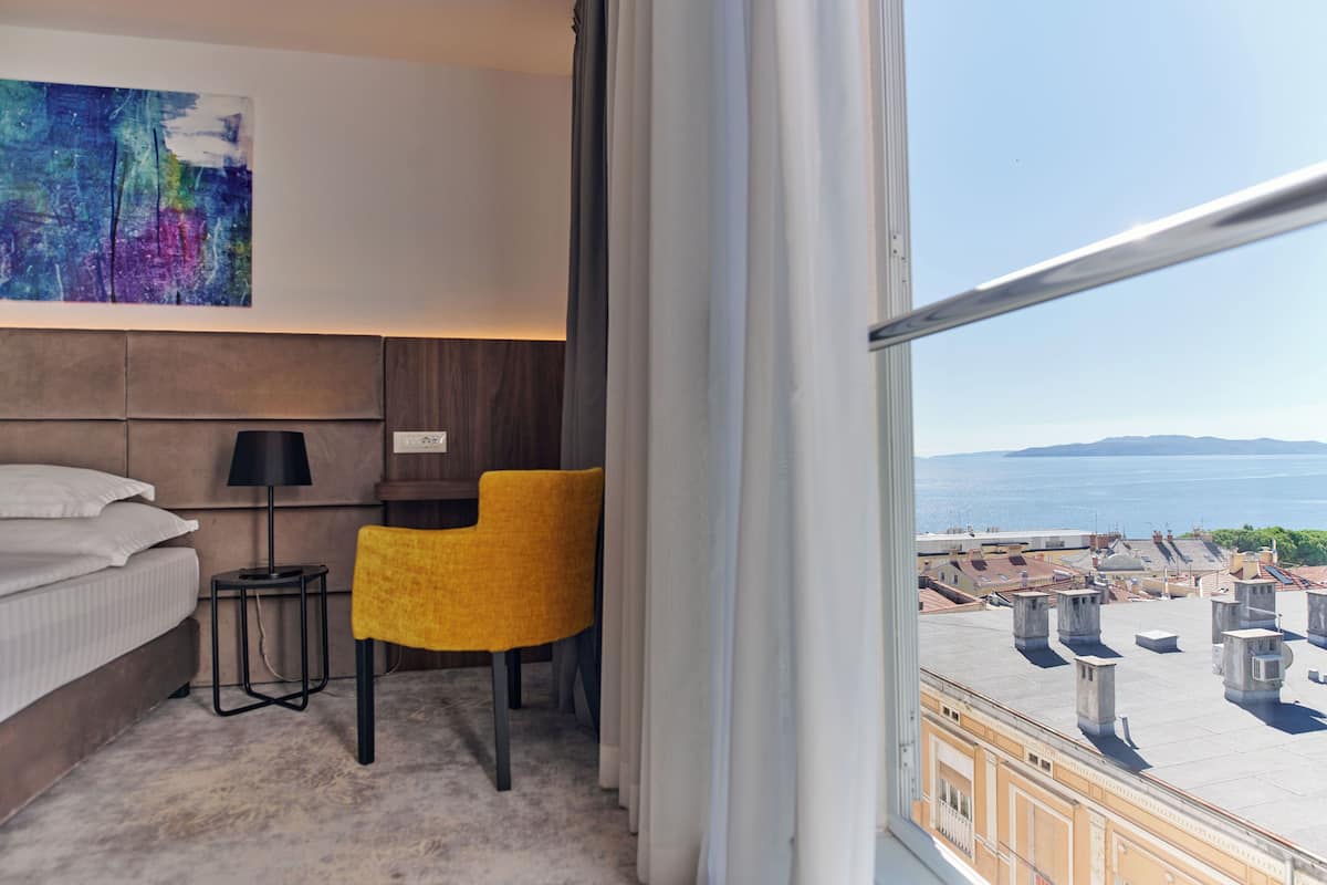 A hotel room with a bed, a desk, a chair, a lamp, and a large window with a view of the city and the sea.