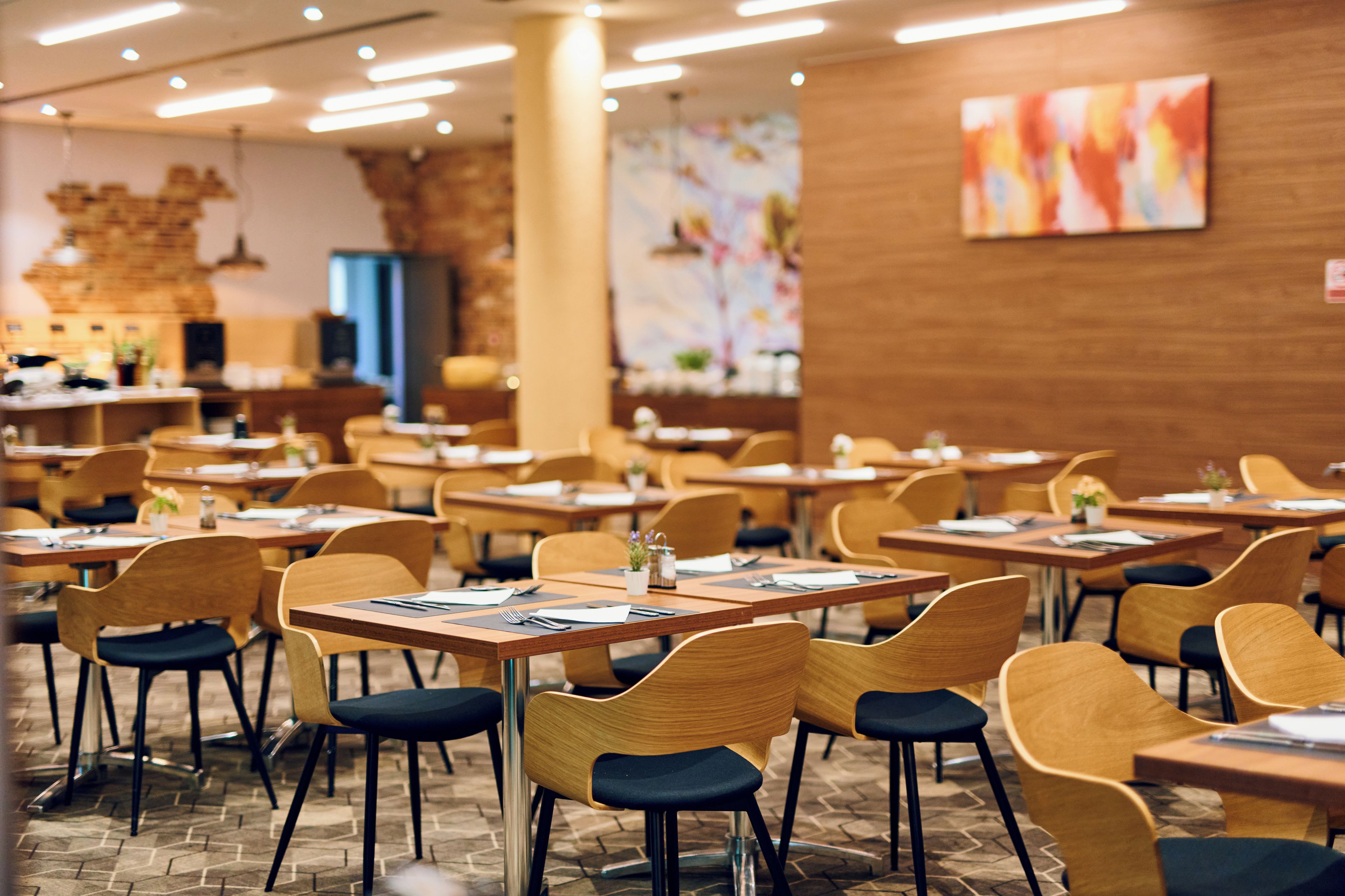 Un restaurant avec des tables et des chaises en bois, un mur en briques et un tableau abstrait aux couleurs vives.