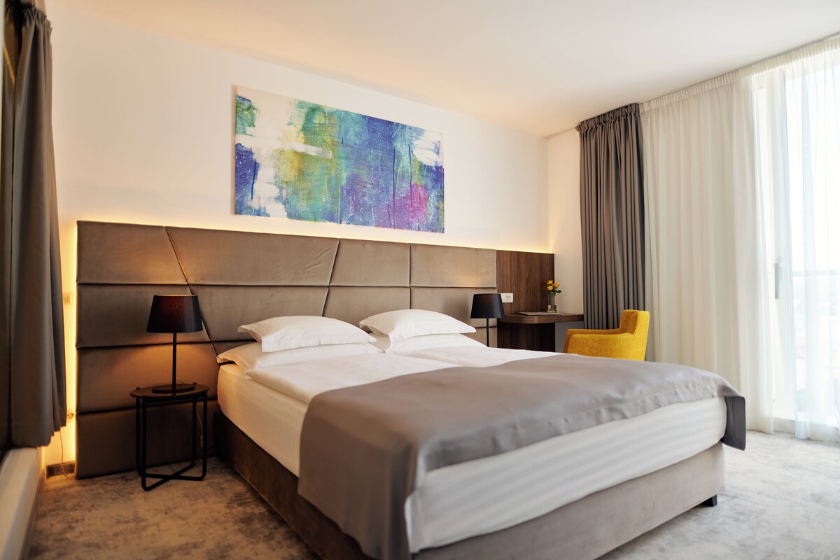 Ein Hotelzimmer mit einem großen Bett, einem Kopfteil, einem Nachttisch mit Lampe, einem Sessel und einem Bild an der Wand.