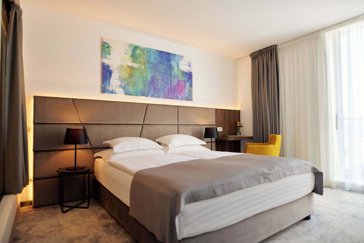 A hotel room with a large bed, a headboard, a side table with a lamp, a chair, and a painting on the wall.