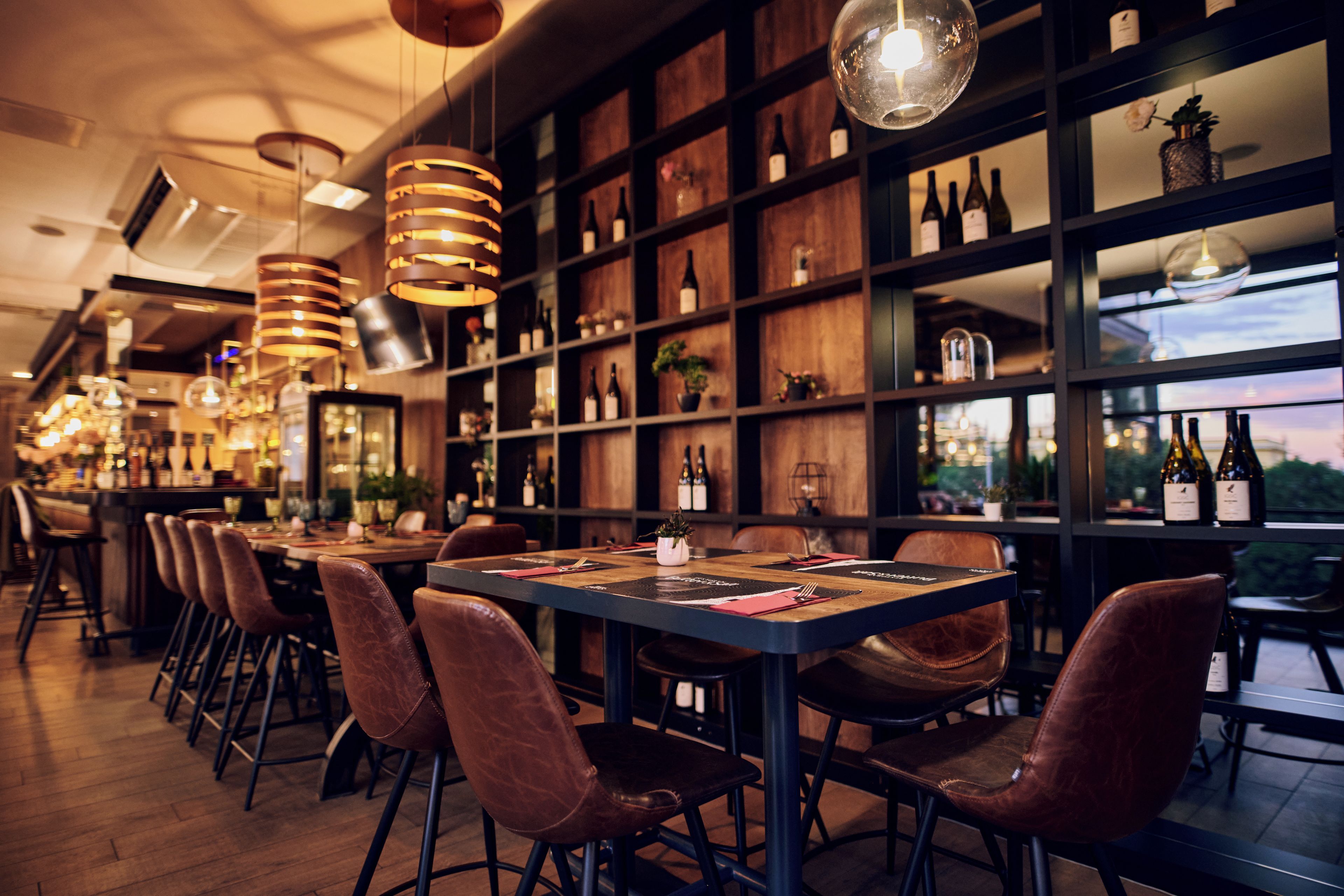 A restaurant with a long counter, multiple chairs, and a wine rack with bottles.