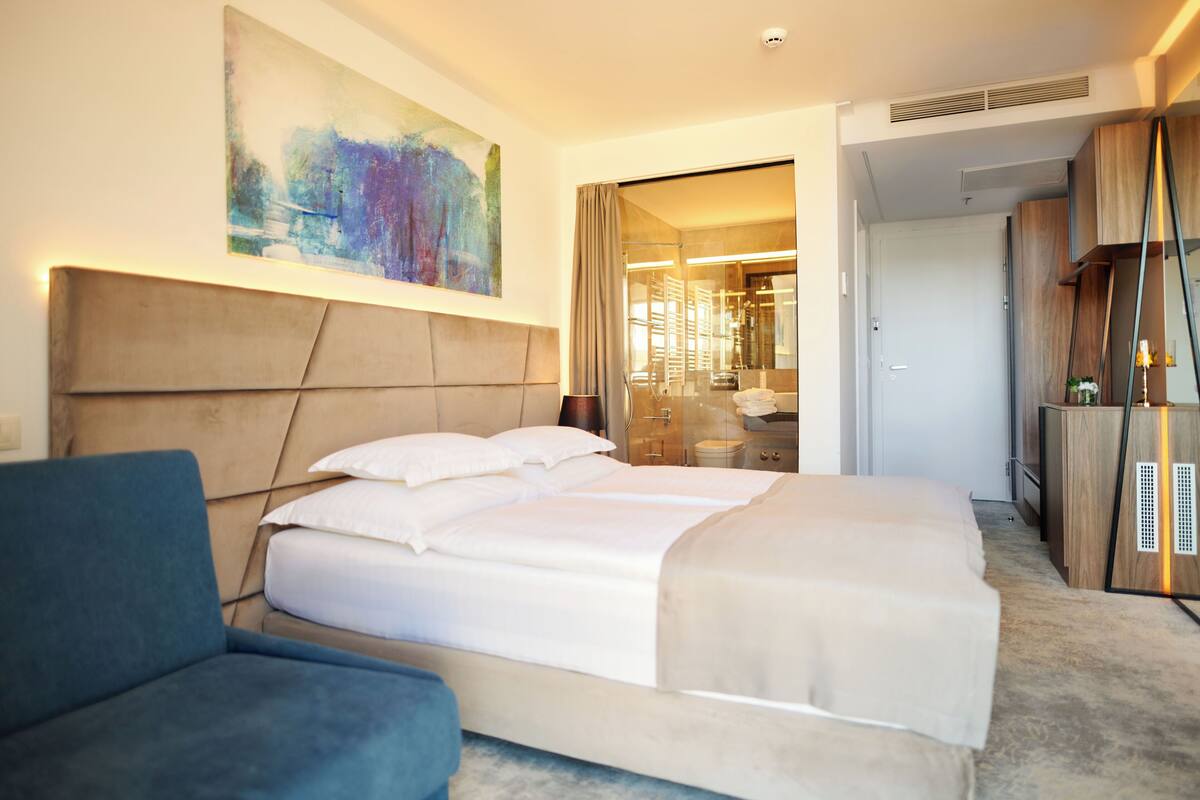 A modern hotel room with a large bed, a blue armchair, and a large abstract painting on the wall.