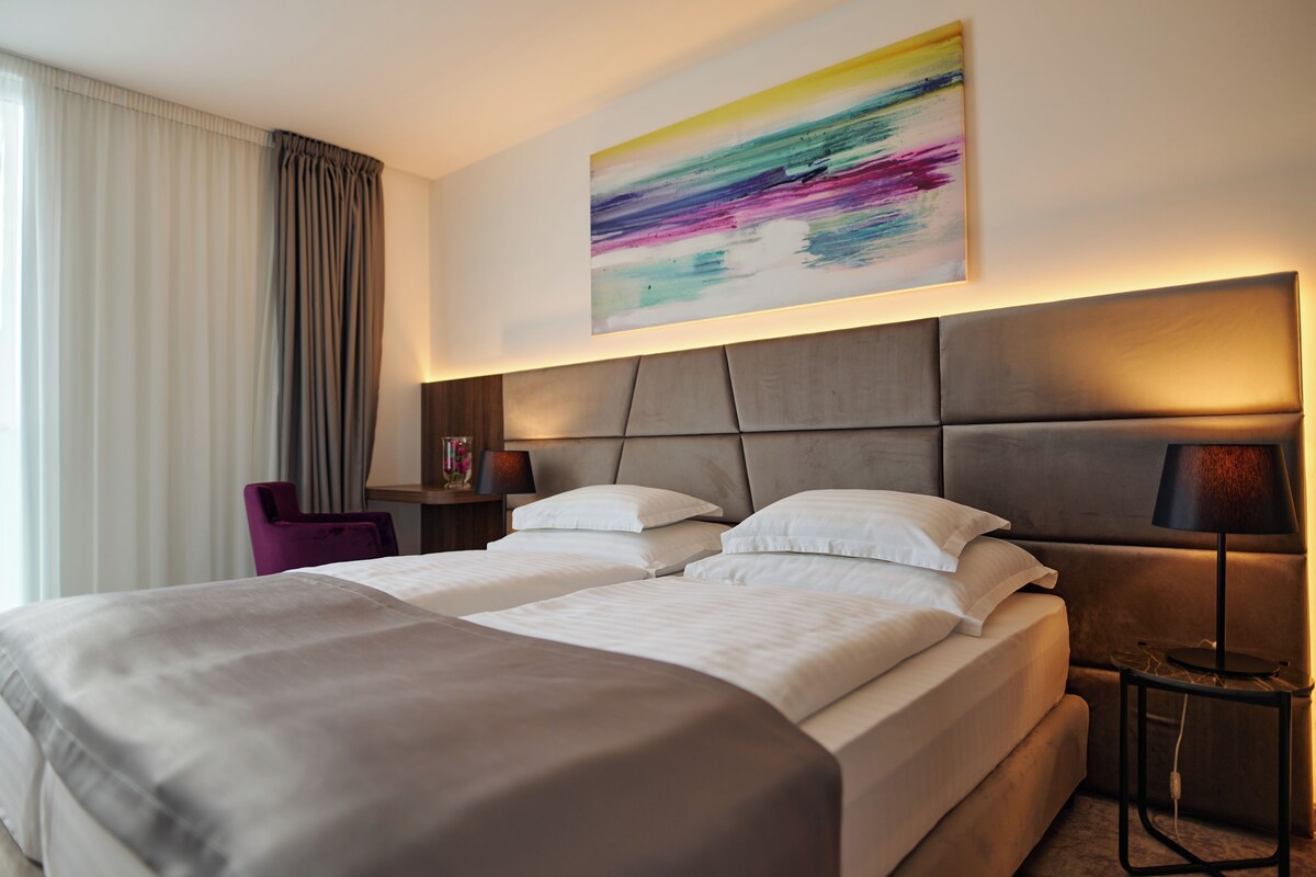 Ein Hotelzimmer mit einem großen Bett, einem Nachttisch mit Lampe, einem Sessel und einem farbenfrohen, abstrakten Gemälde an der Wand.