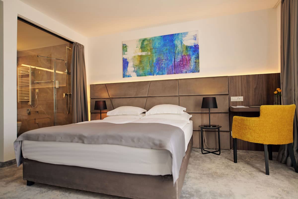 A modern hotel room with a large bed, a desk, a chair, and a colorful abstract painting on the wall.