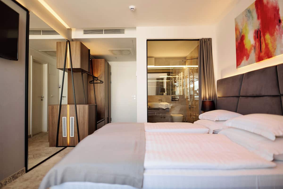 A modern hotel room with a large bed, a wardrobe, a bathroom with a glass shower, and a painting on the wall.