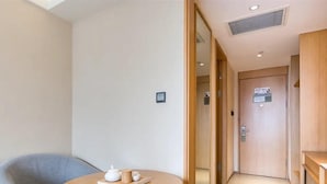 Une chambre d’hôtel moderne avec un canapé gris, une table ronde en bois sur laquelle se trouvent une théière et des tasses, et une porte en bois avec une pancarte.