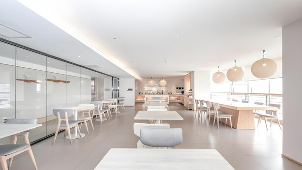 Un espace de restauration moderne et spacieux, avec de longues tables et des chaises, une cuisine en arrière-plan et des luminaires suspendus.