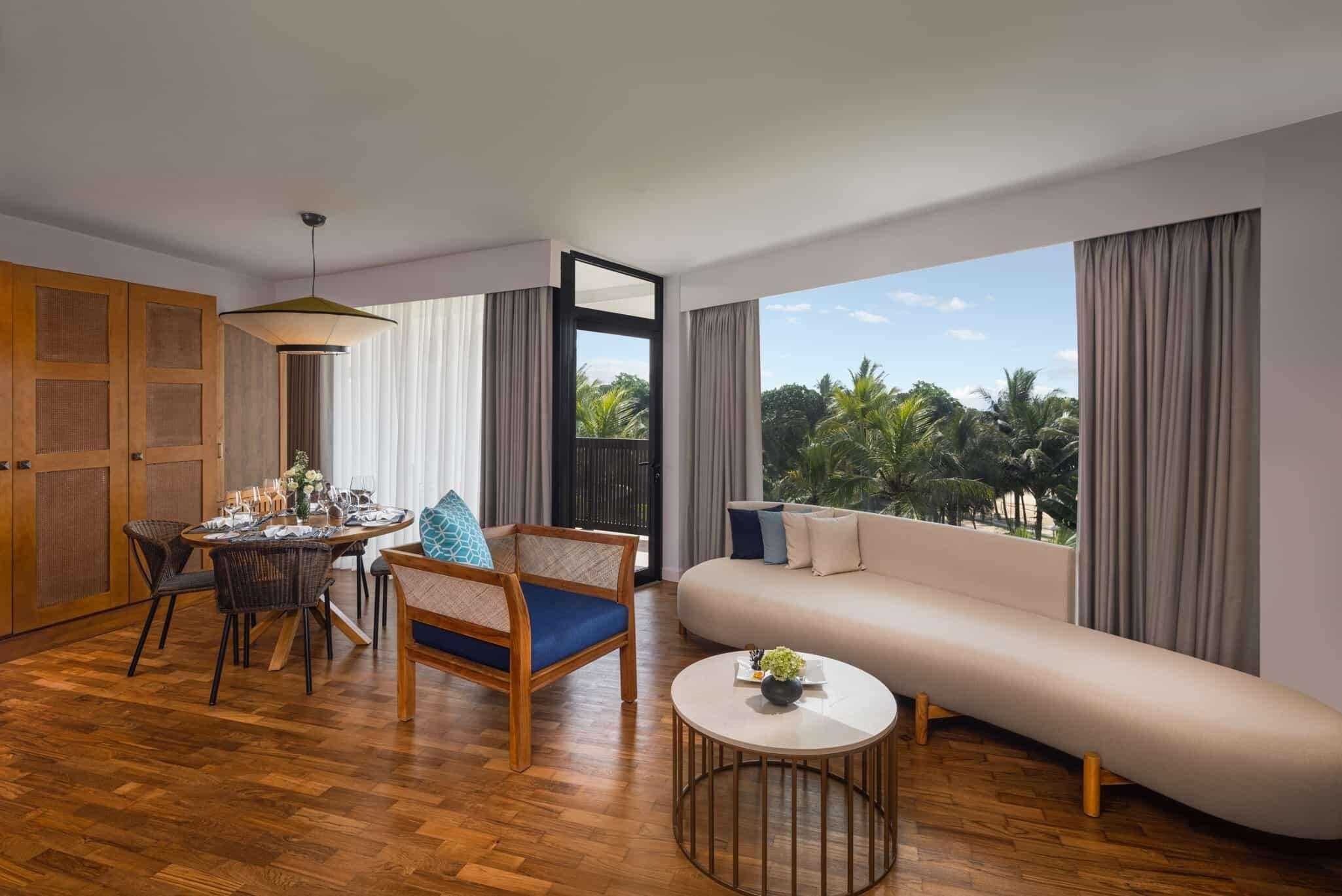 A modern living room with a sofa, a dining table set, and a view of palm trees.