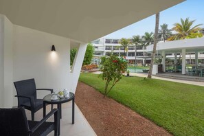 Un área de estar exterior techada con mesa y sillas, un pequeño jardín con un árbol y vistas a un edificio de varios pisos y una piscina.