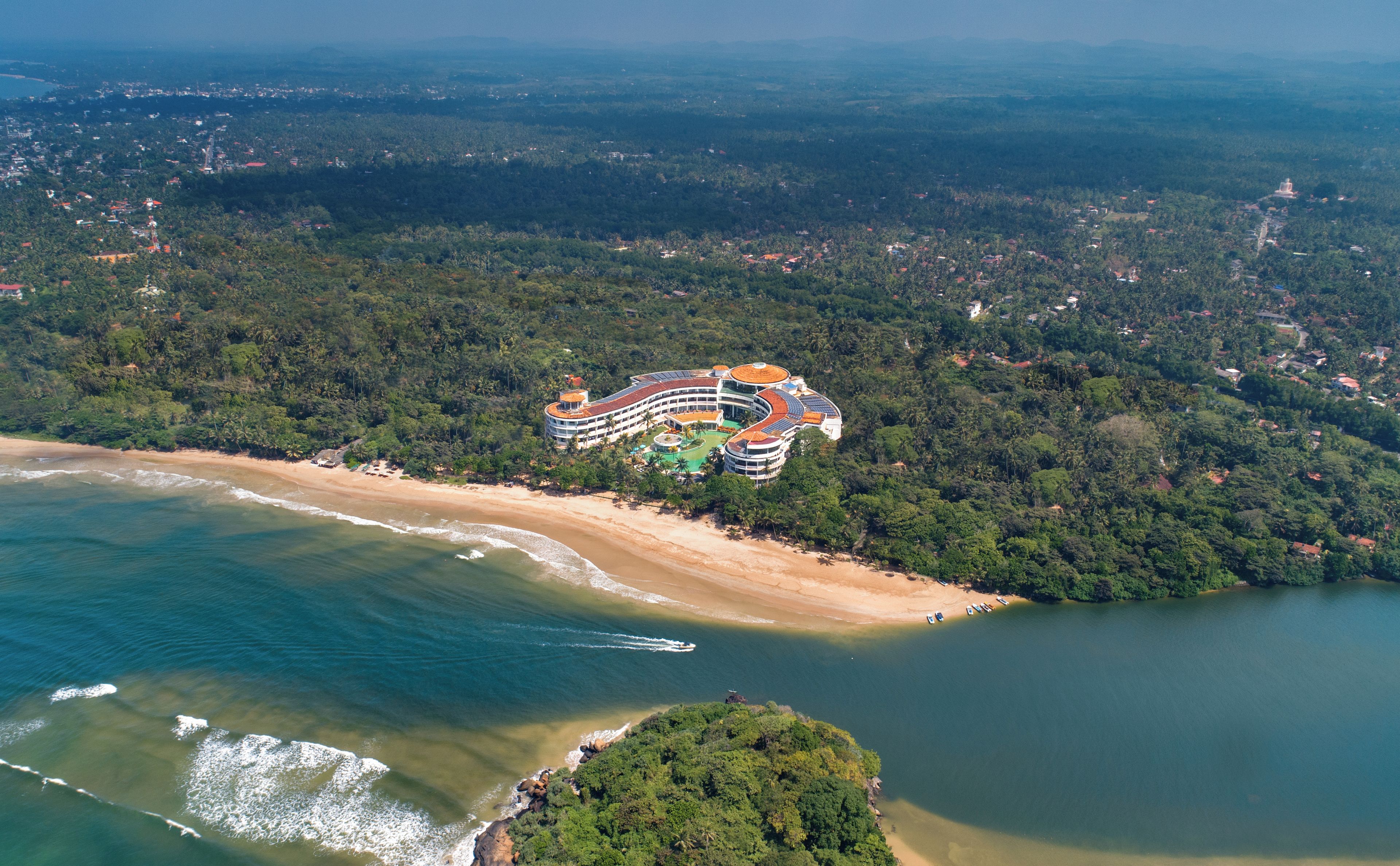 An aerial view of a resort with a beach, a large building with a curved roof, and surrounding forested area.