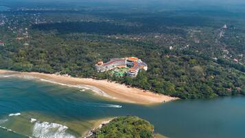 Vista aérea de un complejo turístico con playa, un edificio grande de techo curvo y zonas boscosas circundantes.